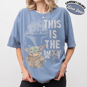 May include: Blue jean-colored t-shirt featuring a graphic of a helmet, a small green creature, and the phrase "THIS IS THE WAY." The shirt also has the words "COMFORT COLORS" and "Blue Jean" in a circular design.