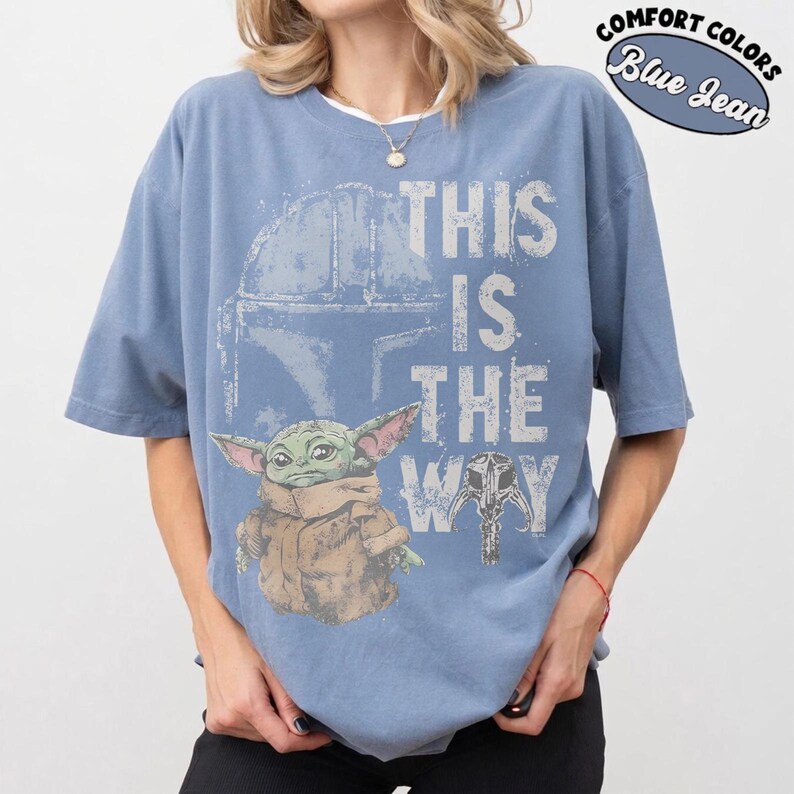 May include: Blue jean-colored t-shirt featuring a graphic of a helmet, a small green creature, and the phrase "THIS IS THE WAY." The shirt also has the words "COMFORT COLORS" and "Blue Jean" in a circular design.