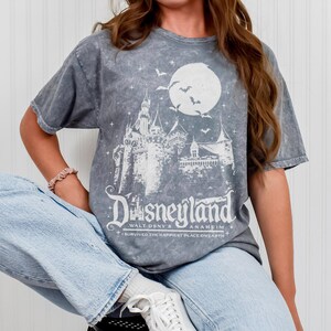 Pode incluir: Camiseta cinza com um gr&aacute;fico branco de um castelo, lua e morcegos, com a palavra "Disneyland" e "Walt Dsny's Anaheim" impressas abaixo. A camisa tamb&eacute;m tem o texto "I survived the happiest place on earth".