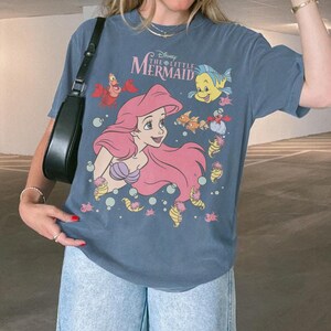 Pode incluir: Camiseta azul com um desenho de Ariel de A Pequena Sereia da Disney. O design inclui Ariel com cabelo rosa, um suti&atilde; de concha roxa e v&aacute;rias criaturas marinhas. O texto "The Little Mermaid" est&aacute; acima da imagem.
