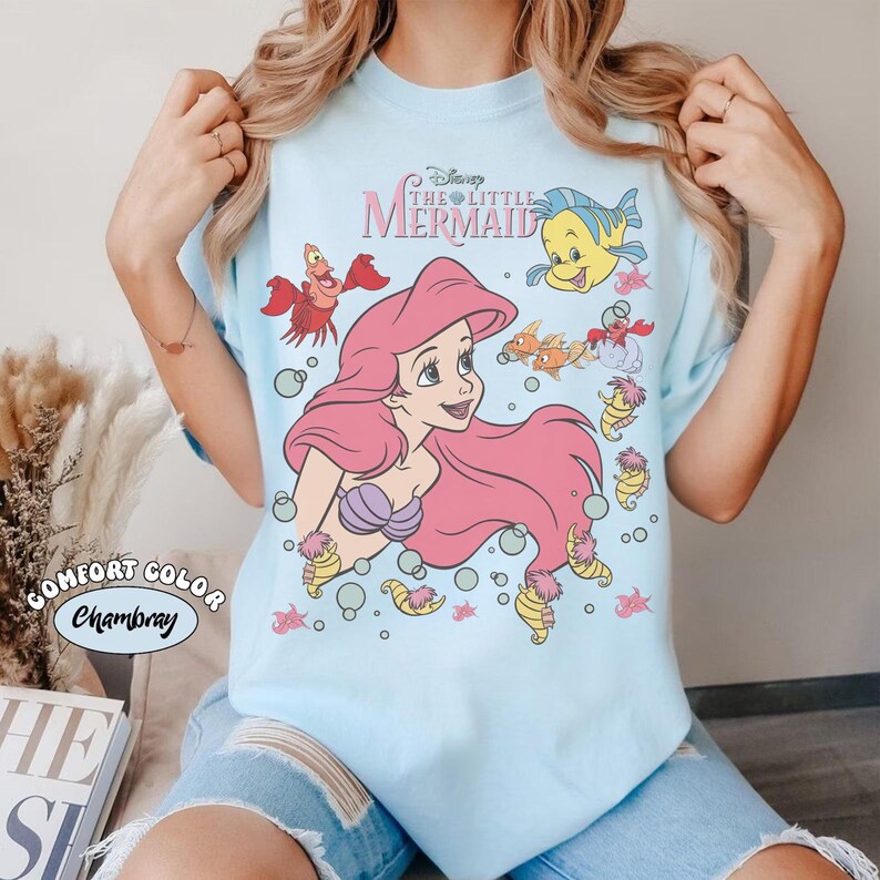 Pode incluir: Camiseta azul clara com um gr&aacute;fico de Ariel de A Pequena Sereia da Disney. Ariel tem cabelo rosa e um top de concha roxa. A camisa tamb&eacute;m apresenta Linguado, Sebasti&atilde;o e outras criaturas marinhas. O texto "Disney The Little Mermaid" est&aacute; acima do gr&aacute;fico.