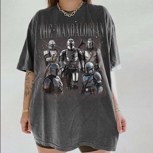 May include: Dark gray t-shirt featuring the text "THE MANDALORIAN" above a graphic of several Mandalorian characters in armor. The shirt has a vintage, worn look.