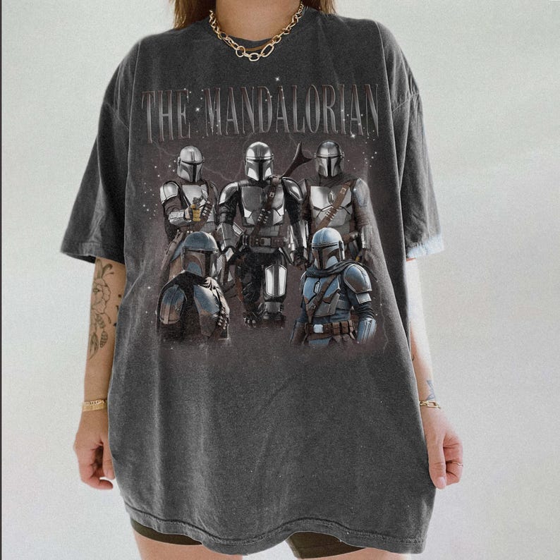May include: Dark gray t-shirt featuring the text "THE MANDALORIAN" above a graphic of several Mandalorian characters in armor. The shirt has a vintage, worn look.