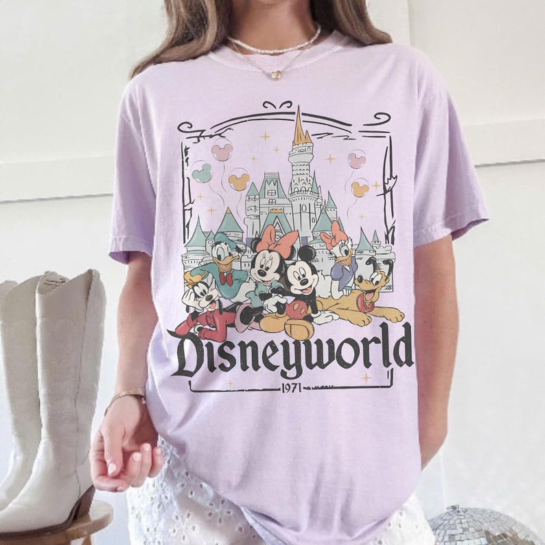 May include: A light purple t-shirt featuring a Disneyworld graphic. The graphic includes Disney characters like Mickey and Minnie Mouse, Donald Duck, and Pluto, with a castle backdrop and the text "Disneyworld 1971."