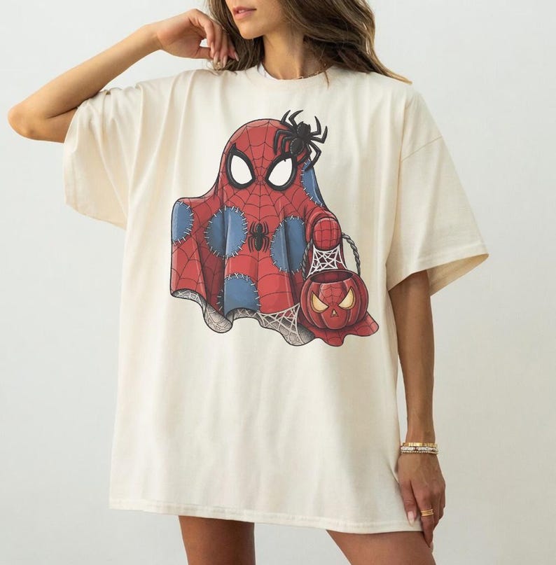 May include: Cream-colored t-shirt featuring a Halloween-themed graphic. The design depicts a ghost in a Spider-Man costume holding a pumpkin-shaped candy bucket. The ghost is red and blue with a spider web design.