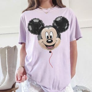 Puede incluir: Camiseta lila claro con un dise&ntilde;o de globo de Mickey Mouse. El globo tiene orejas negras, una cara beige y una cuerda roja. La camiseta es de estilo casual.