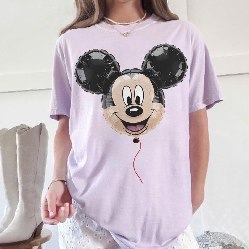 Puede incluir: Camiseta lila claro con un dise&ntilde;o de globo de Mickey Mouse. El globo tiene orejas negras, una cara beige y una cuerda roja. La camiseta es de estilo casual.
