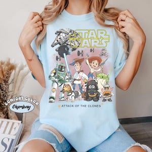 May include: Pale blue t-shirt with a graphic of Star Wars characters and the text "STAR WARS" in yellow. The shirt also includes the text "ATTACK OF THE CLONES". The shirt is a Comfort Color Chambray.