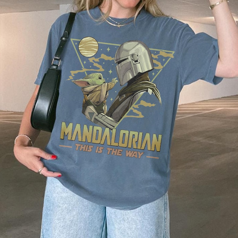 May include: Blue t-shirt featuring a graphic of The Mandalorian and Grogu, with the text "MANDALORIAN THIS IS THE WAY". The design includes a triangle with a moon and stars. The shirt is made of a soft material.