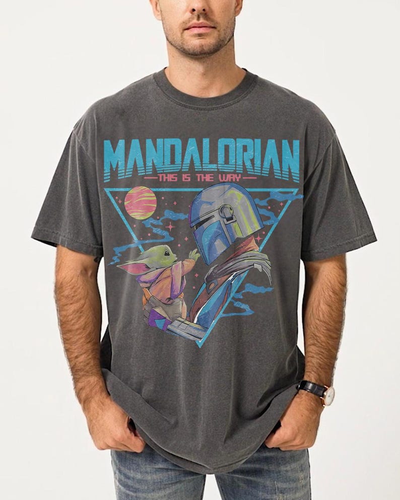 May include: Dark gray t-shirt featuring a graphic of The Mandalorian and Grogu, with the text "MANDALORIAN - THIS IS THE WAY" in blue. The design includes a triangle with a planet and stars.