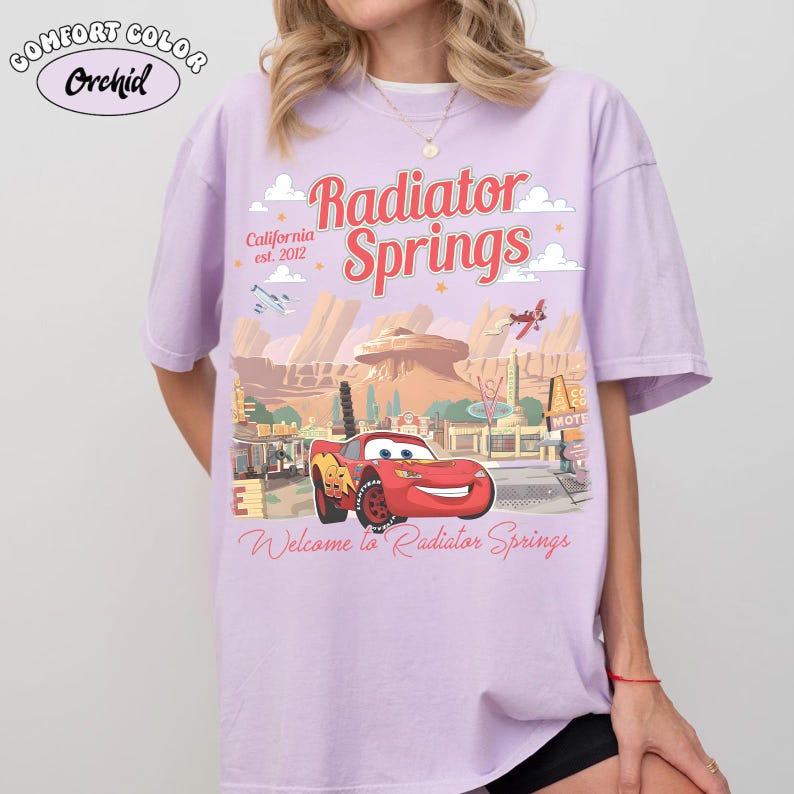 May include: A light purple t-shirt featuring a graphic of the town of Radiator Springs from the movie Cars. The design includes the text "Radiator Springs" in red, the text "California est. 2012", and a cartoon image of the character Lightning McQueen.