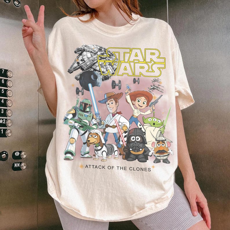 May include: Cream-coloured t-shirt with a vibrant graphic of Star Wars characters and the yellow text "STAR WARS". The design includes the Millennium Falcon, Darth Vader, and other characters from the film. The text "ATTACK OF THE CLONES" is also visible.