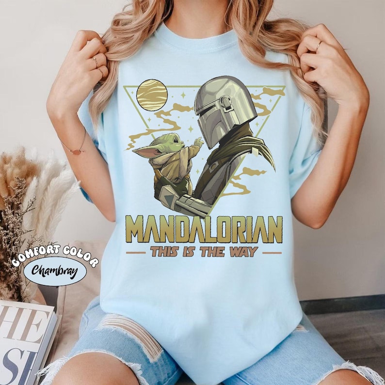 May include: Light blue t-shirt featuring a graphic of The Mandalorian and Grogu. The design includes the text "MANDALORIAN - THIS IS THE WAY" in gold lettering. The shirt is a Comfort Color Chambray.