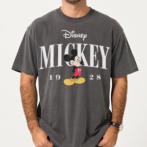 May include: Charcoal grey t-shirt with the Disney logo and the word "MICKEY" in white. Below the word "MICKEY" is a cartoon image of Mickey Mouse and the numbers "19 28".