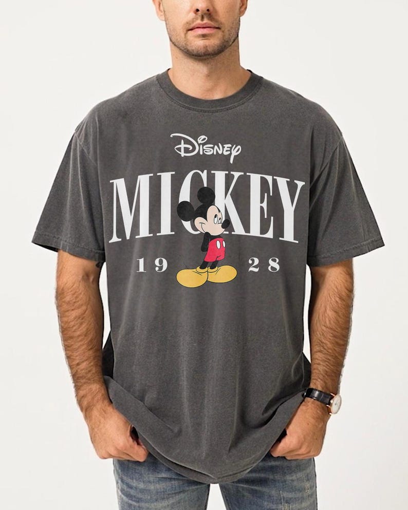 May include: Charcoal grey t-shirt with the Disney logo and the word "MICKEY" in white. Below the word "MICKEY" is a cartoon image of Mickey Mouse and the numbers "19 28".