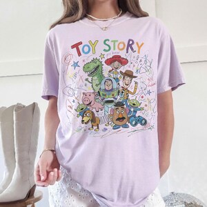 May include: A light purple t-shirt featuring a colorful graphic of characters from the animated film "Toy Story." The design includes the words "Toy Story" in a rainbow font, along with drawings of Woody, Buzz Lightyear, and other characters.