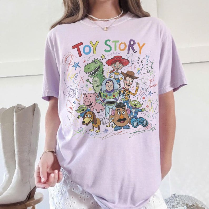 May include: A light purple t-shirt featuring a colorful graphic of characters from the animated film "Toy Story." The design includes the words "Toy Story" in a rainbow font, along with drawings of Woody, Buzz Lightyear, and other characters.