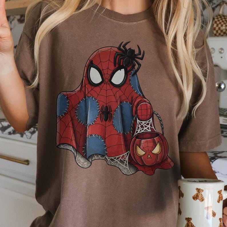 May include: Brown t-shirt featuring a Spiderman-themed ghost design with a spider on top. The ghost is red with a spiderweb pattern and blue patches. A pumpkin-shaped basket with a spiderweb design is also visible.