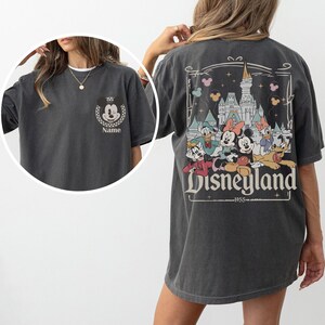 May include: Dark gray t-shirt featuring Mickey Mouse on the front and a Disneyland graphic on the back. The front design includes Mickey's face with the word "Name" below. The back graphic showcases Disney characters and the Disneyland logo.