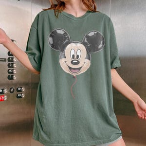 Puede incluir: Camiseta verde con un dise&ntilde;o de globo de Mickey Mouse. El globo tiene orejas negras y una cara sonriente, con una cuerda roja. La camiseta es de corte holgado.