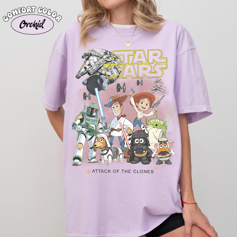 May include: A lilac t-shirt with a Star Wars design, showcasing characters and the text "ATTACK OF THE CLONES." The graphic includes a spaceship, a lightsaber, and the Star Wars logo in yellow.