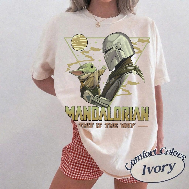 May include: Ivory-colored t-shirt featuring a graphic of The Mandalorian and Grogu. The design includes the text "MANDALORIAN" and "THIS IS THE WAY". The shirt is a Comfort Colors brand.