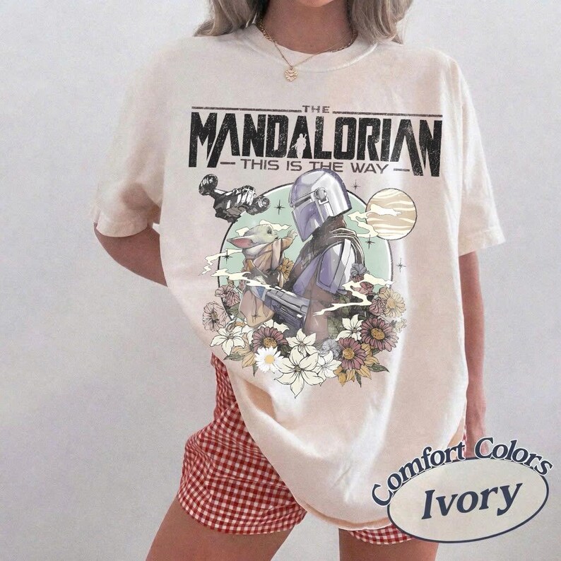 May include: Ivory-colored t-shirt featuring a graphic of The Mandalorian and Grogu surrounded by flowers, with the text "THE MANDALORIAN THIS IS THE WAY." The shirt is a Comfort Colors brand.