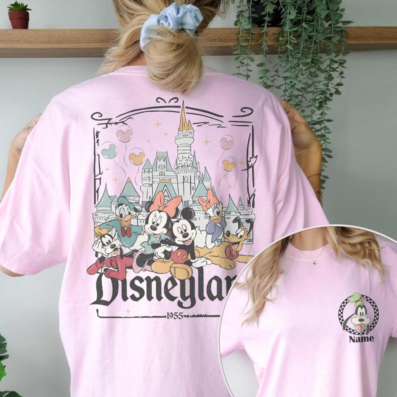 May include: Pink t-shirt featuring a Disneyland graphic with Mickey Mouse, Minnie Mouse, Donald Duck, Daisy Duck, Goofy, and Pluto. The graphic includes a castle and the text "Disneyland 1955". A smaller image of Goofy is on the front.