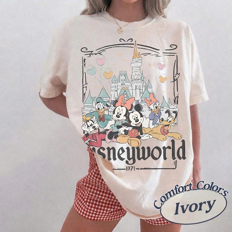 May include: Ivory-colored t-shirt featuring a Disney World graphic with Mickey, Minnie, Donald, Daisy, Goofy, and Pluto. The graphic includes a castle, balloons, and the text "Disneyworld 1971".
