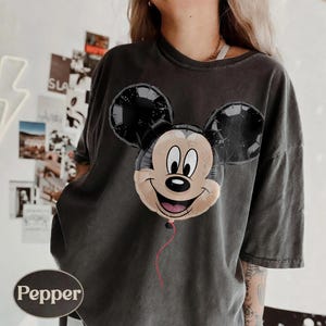 Puede incluir: Camiseta gris oscuro con un dise&ntilde;o de globo de Mickey Mouse. El globo tiene orejas negras y una cara sonriente con una cuerda roja. La camiseta tiene un corte holgado y un aspecto vintage.