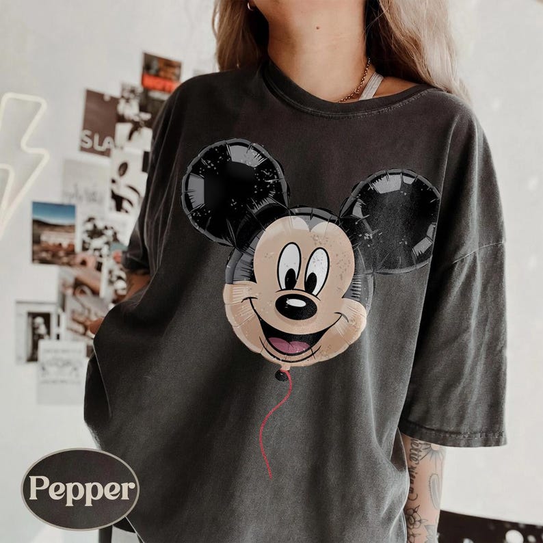 Puede incluir: Camiseta gris oscuro con un dise&ntilde;o de globo de Mickey Mouse. El globo tiene orejas negras y una cara sonriente con una cuerda roja. La camiseta tiene un corte holgado y un aspecto vintage.