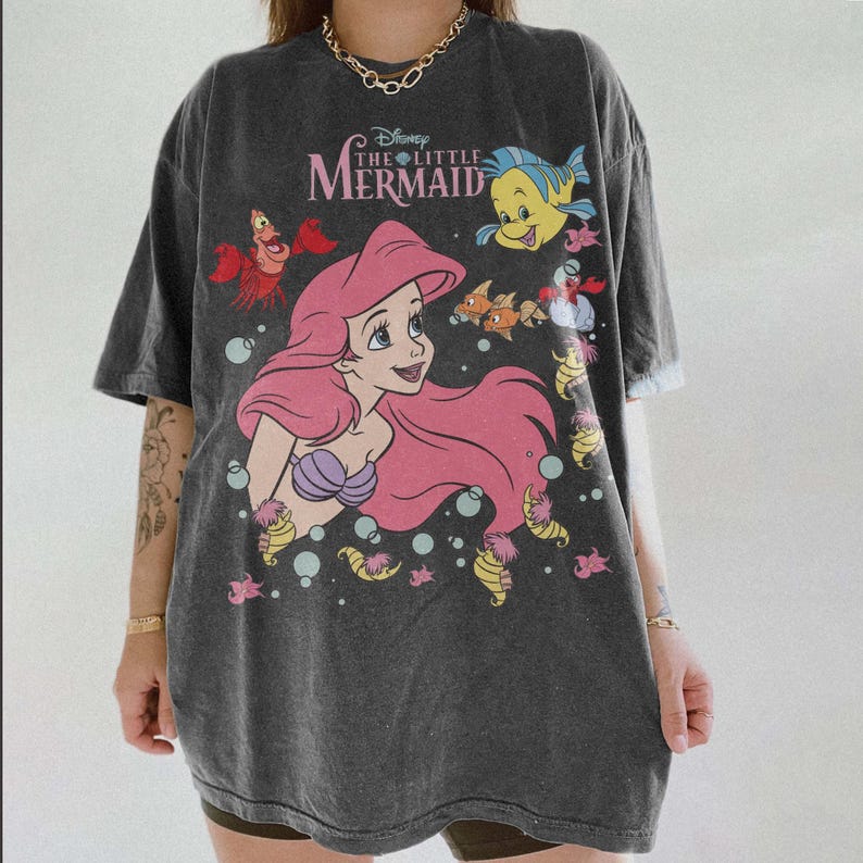 Pode incluir: Camiseta cinza escuro com um gr&aacute;fico de Ariel de A Pequena Sereia da Disney. O design inclui Ariel com cabelo rosa, um suti&atilde; de concha roxa e v&aacute;rias criaturas marinhas. O texto "The Little Mermaid" &eacute; impresso acima do gr&aacute;fico.