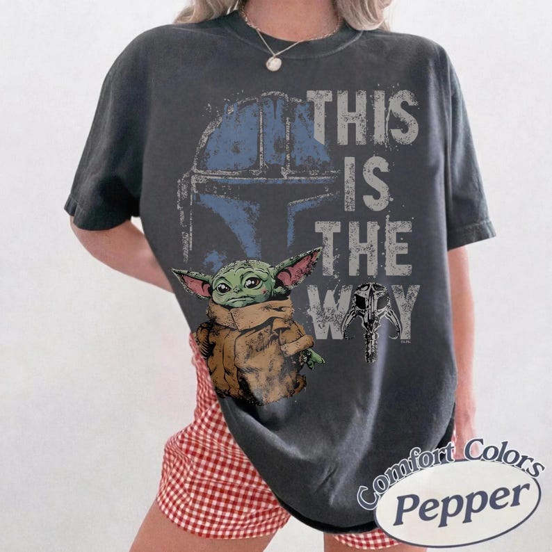May include: Dark gray t-shirt featuring a graphic of a Mandalorian helmet, Baby Yoda, and the phrase "THIS IS THE WAY" in white and blue. The shirt is a Comfort Colors brand.