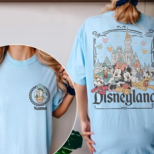 May include: Light blue t-shirt featuring a Donald Duck graphic on the front and a Disneyland graphic on the back. The Disneyland graphic includes Mickey and Minnie Mouse, and the text "Disneyland 1955".