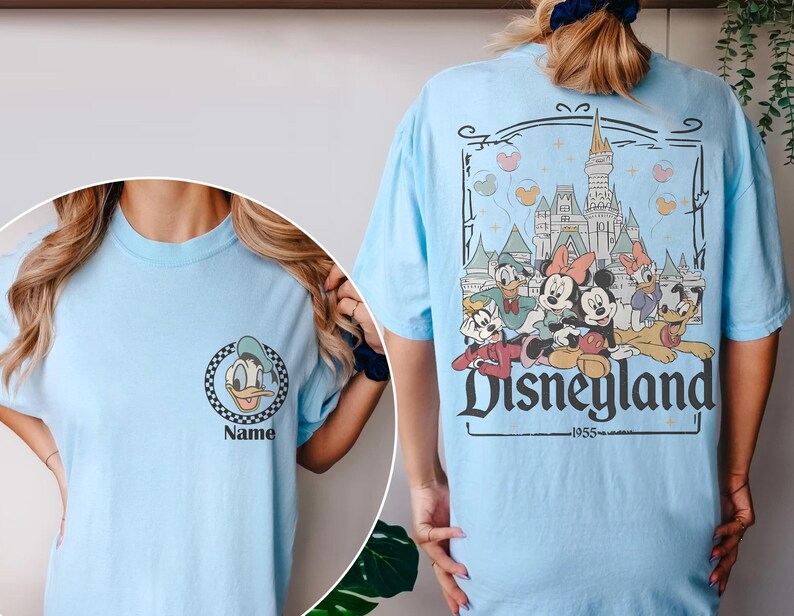 May include: Light blue t-shirt featuring a Donald Duck graphic on the front and a Disneyland graphic on the back. The Disneyland graphic includes Mickey and Minnie Mouse, and the text "Disneyland 1955".
