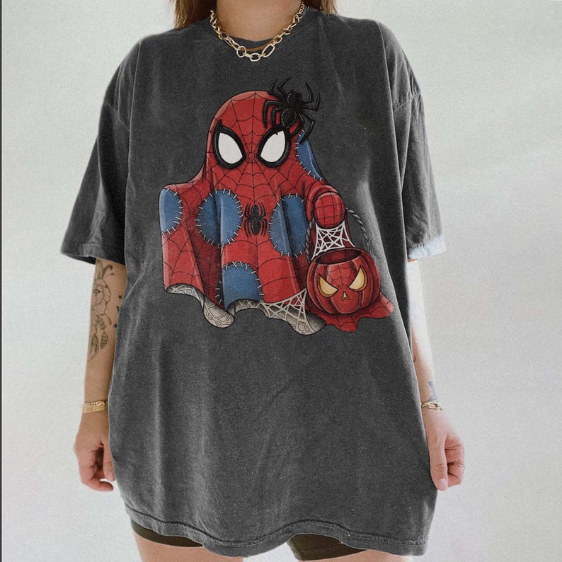May include: Dark gray t-shirt featuring a Halloween-themed graphic. The design depicts a ghost in a Spider-Man costume holding a jack-o'-lantern. The ghost is red and blue with a black spider on top. The shirt is oversized.