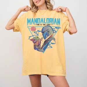 May include: Yellow t-shirt featuring a graphic of The Mandalorian and Grogu, with the text "MANDALORIAN - THIS IS THE WAY" in blue. The design includes a triangle, a planet, and blue accents. The shirt is a comfortable fit.
