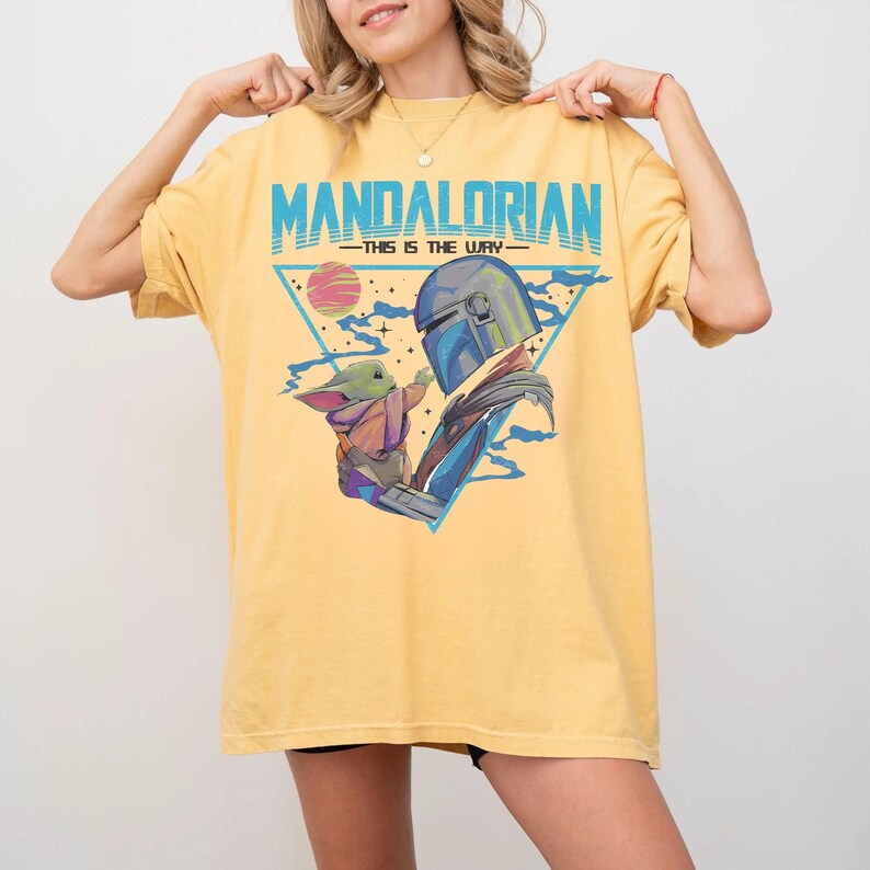 May include: Yellow t-shirt featuring a graphic of The Mandalorian and Grogu, with the text "MANDALORIAN - THIS IS THE WAY" in blue. The design includes a triangle, a planet, and blue accents. The shirt is a comfortable fit.