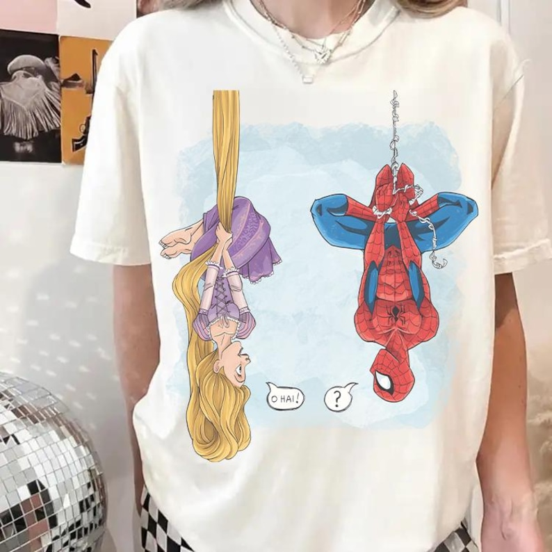 Op de afbeelding: Cr&egrave;mekleurig T-shirt met een cartoonillustratie van Rapunzel en Spider-Man die ondersteboven hangen. Rapunzel houdt haar lange blonde haar vast en Spider-Man hangt aan een web. Spraakballonnen zeggen "O HAI!" en "?".