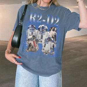 May include: Blue t-shirt featuring a graphic of several R2-D2 droids from Star Wars. The text "R2-D2" is printed above the droids in a gradient blue color. The shirt is a casual style.