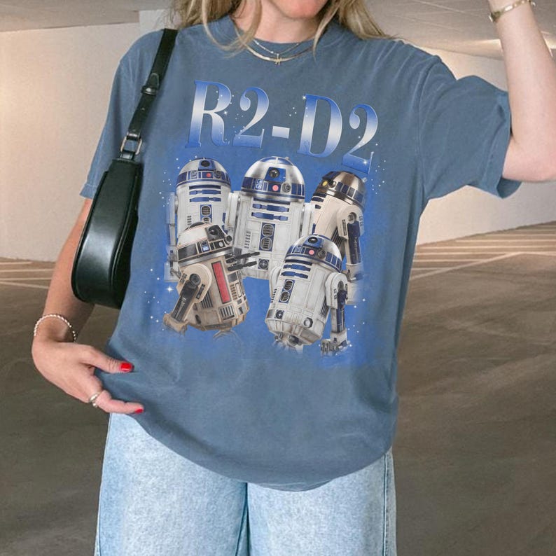 May include: Blue t-shirt featuring a graphic of several R2-D2 droids from Star Wars. The text "R2-D2" is printed above the droids in a gradient blue color. The shirt is a casual style.