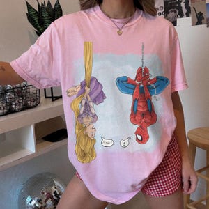 Op de afbeelding: Zachtroze T-shirt met een cartoon van Rapunzel en Spider-Man die ondersteboven hangen. Rapunzel hangt aan haar haar en Spider-Man hangt aan een web. Het shirt heeft de tekst "O Hai!" en een vraagteken.