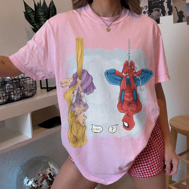 Op de afbeelding: Zachtroze T-shirt met een cartoon van Rapunzel en Spider-Man die ondersteboven hangen. Rapunzel hangt aan haar haar en Spider-Man hangt aan een web. Het shirt heeft de tekst "O Hai!" en een vraagteken.