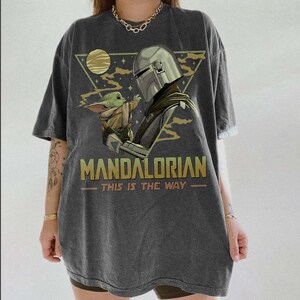May include: Dark gray t-shirt featuring a graphic of The Mandalorian and Grogu. The design includes the text "MANDALORIAN" and "THIS IS THE WAY" in gold lettering, with a triangular background and a moon.