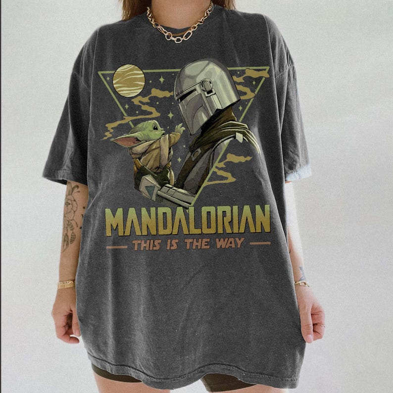 May include: Dark gray t-shirt featuring a graphic of The Mandalorian and Grogu. The design includes the text "MANDALORIAN" and "THIS IS THE WAY" in gold lettering, with a triangular background and a moon.