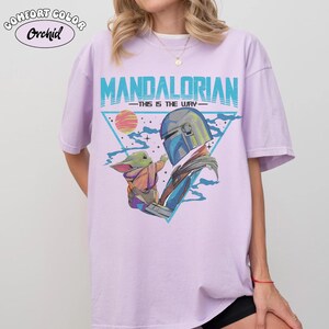 May include: Light purple t-shirt with a graphic of The Mandalorian and Grogu. The design features the text "MANDALORIAN" in teal, with "THIS IS THE WAY" underneath. The image includes a triangle, a sun, and Grogu reaching towards The Mandalorian.
