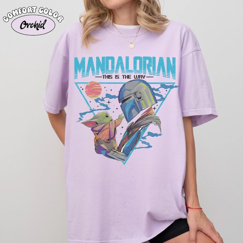 May include: Light purple t-shirt with a graphic of The Mandalorian and Grogu. The design features the text "MANDALORIAN" in teal, with "THIS IS THE WAY" underneath. The image includes a triangle, a sun, and Grogu reaching towards The Mandalorian.