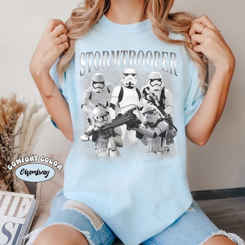 May include: Light blue t-shirt featuring a graphic of Stormtroopers from Star Wars. The word "STORMTROOPER" is printed across the top in silver lettering. The shirt is a Comfort Color Chambray.