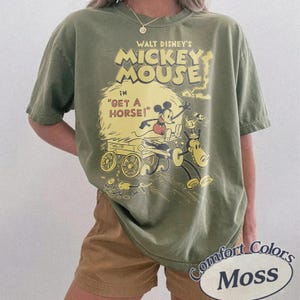 May include: Olive green t-shirt featuring a vintage Mickey Mouse graphic from "Get a Horse!" The design includes Mickey Mouse, a horse, and a cart in yellow and red. The shirt is labeled "Comfort Colors Moss".