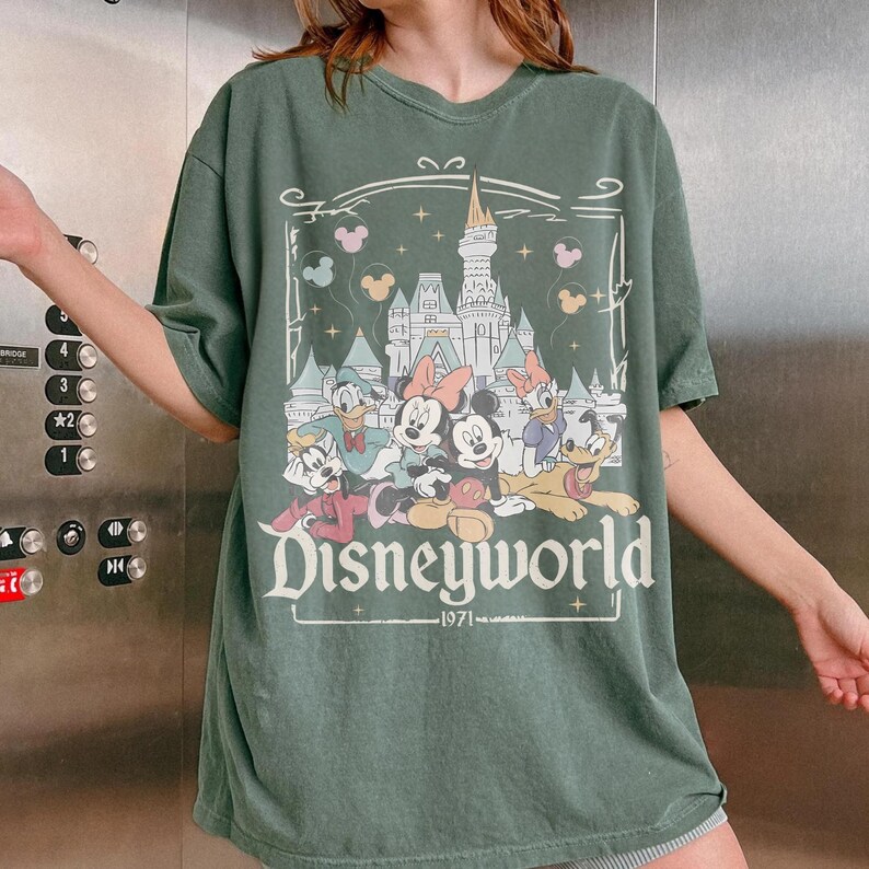 May include: Green t-shirt featuring a Disneyworld graphic with Mickey Mouse, Minnie Mouse, Donald Duck, and Goofy. The graphic includes a castle, stars, and the text "Disneyworld 1971". The shirt has a crew neck and short sleeves.